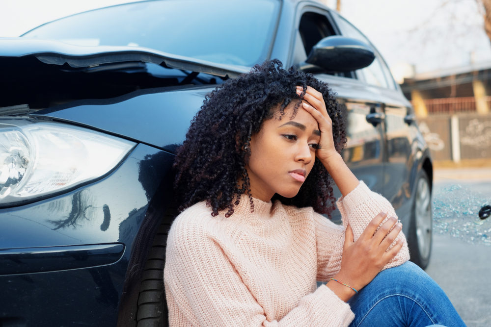 When your car accident claim exceeds insurance policy limits Black woman leaning against a car holding her head after a crash