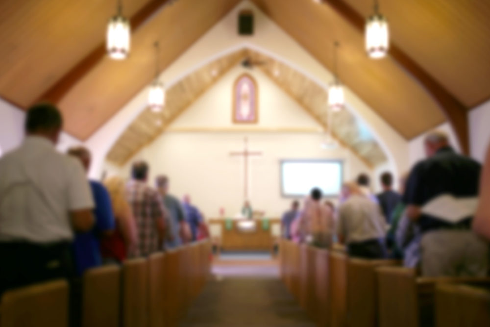El Gran Rey Jesus Pastor, Abner Vazquez, Accused of Sexually Assaulting Woman A blurred photo of the inside of a church sanctuary that is filled with people in the pews, and the pastor stands under a large cross at the altar.