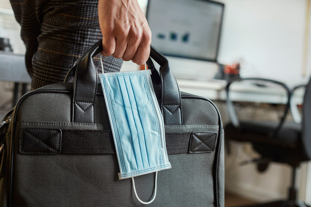 Guide to COVID-19 Vaccines In the Workplace closeup of a young man in an office holding a briefcase and a surgical mask in his hand