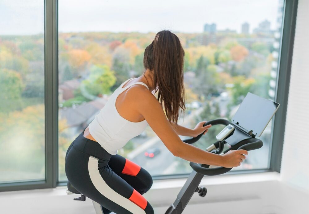 Peloton Recalls 2.2 Million Exercise Bikes Due to Safety Hazard woman exercising at home indoor cycling with online class on screen.