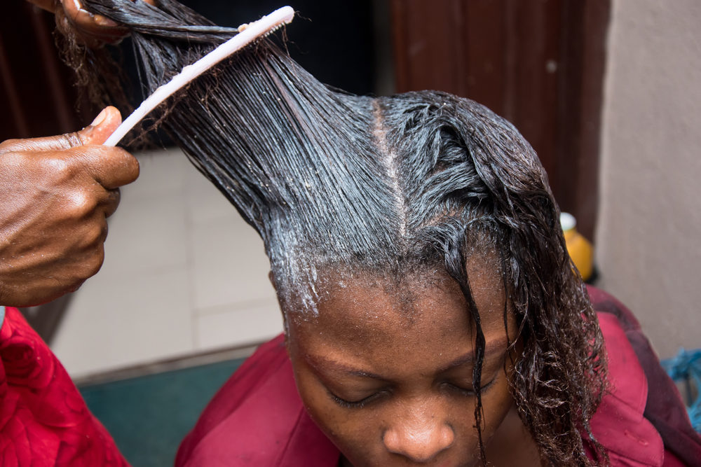 Arizona Woman Undergoes Partial Hysterectomy for Uterine Fibroids—Blames Hair-Relaxing Products Hairdresser relaxing the hair on an African woman head and also using comb to stretch and apply the relaxer cream through the hair