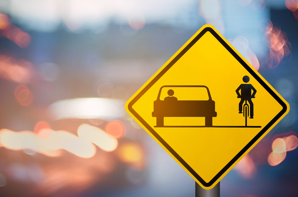 yellow warning sign for  a shared roadway with a car driving alongside a bicyclist