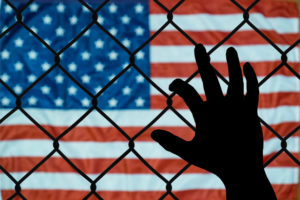 Silhouetted hand grasping a chain-link fence with American flag on the other side